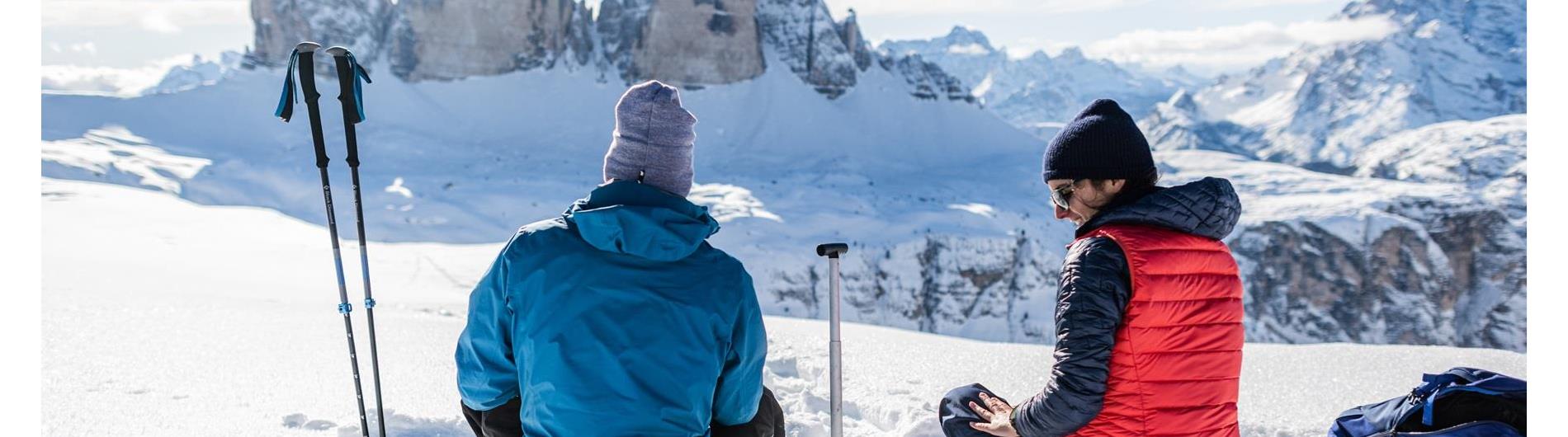 Due persone che fanno escursione con le ciaspole si fermano per una breve pausa e ammirano le Tre Cime di Lavaredo