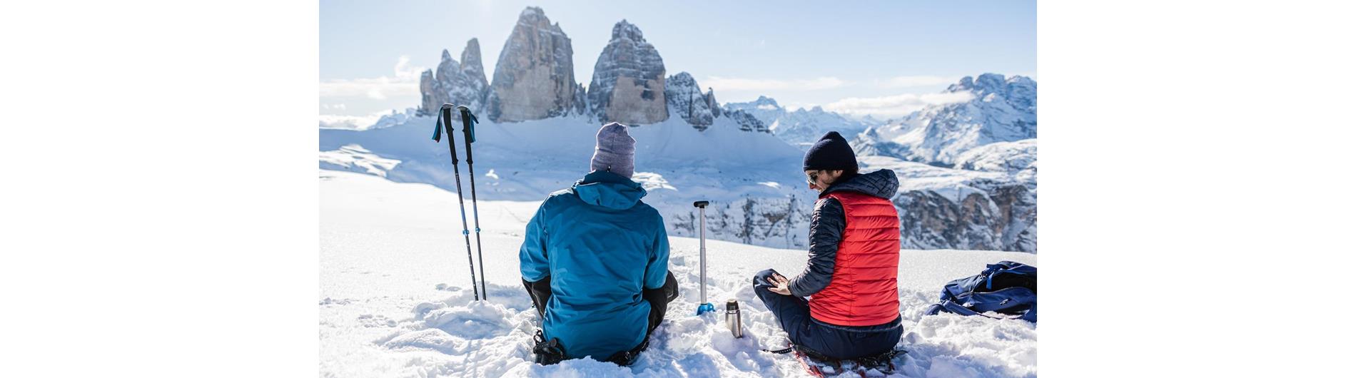 Due persone che fanno escursione con le ciaspole si fermano per una breve pausa e ammirano le Tre Cime di Lavaredo